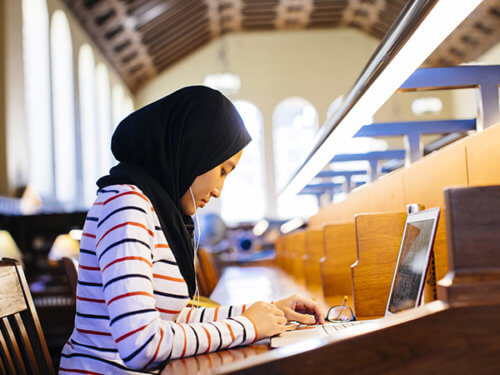 person wearing a headscarf and earbuds seated at a library carrel desk using a laptop computer 