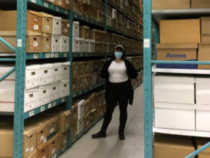 photo of a woman standing amongst archival boxes on shelves 