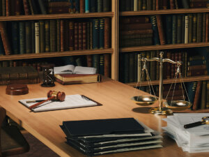 desk topped with legal papers, a gavel, and scales with a bookcase in the background