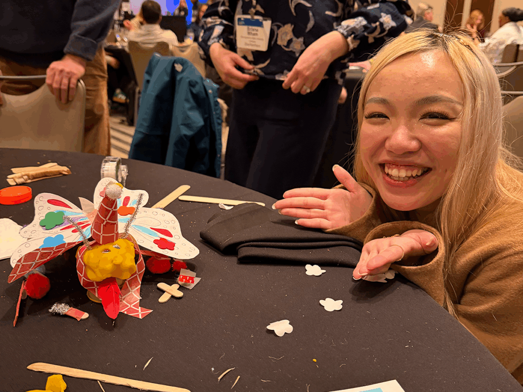 The author poses beside a colorful craft “innovation creature” made from pipe cleaners, paper shapes, and other materials during the Dinn-o-vate activity.