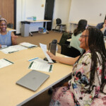 photo of 4 adults seated at tables in a classroom, interacting, and smiling